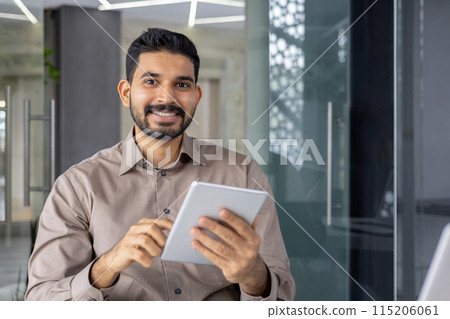 Confident young professional man holding a tablet, smiling and looking approachable in a bright, modern office environment. He appears friendly and tech-savvy. 115206061