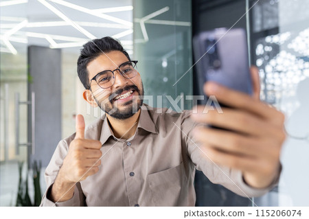 Playful latino guy in eyeglasses smiling widely and showing thumb up gesture while taking selfie on frontal camera of telephone. Relaxed person having fun during working day in company office. Playful latino guy in eyeglasses smiling widely and showing thumb up gesture while taking selfie on frontal camera of telephone. Relaxed person having fun during working day in company office. 115206074