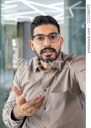 First person view of brunette man in cotton shirt wearing stylish eyeglasses and gesturing to camera during video meeting. Arab company boss sharing important information with employers of department. First person view of brunette man in cotton shirt wearing stylish eyeglasses and gesturing to camera during video meeting. Arab company boss sharing important information with employers of department. 115206075