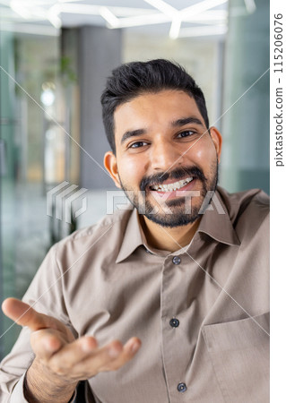 A cheerful man with a beard in a casual shirt gestures while engaging in a video call. The indoor environment is modern and well-lit, highlighting a welcoming video chat experience. A cheerful man with a beard in a casual shirt gestures while engaging in a video call. The indoor environment is modern and well-lit, highlighting a welcoming video chat experience. 115206076