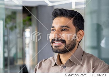 A portrait of a confident young man with a beard smiling warmly in a well-lit, contemporary office environment. The image conveys a sense of happiness and professional success. A portrait of a confident young man with a beard smiling warmly in a well-lit, contemporary office environment. The image conveys a sense of happiness and professional success. 115206341