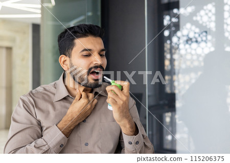 Young man applying throat spray for sore throat relief in a modern office environment. Concept of healthcare, illness, and self-care at the workplace. Young man applying throat spray for sore throat relief in a modern office environment. Concept of healthcare, illness, and self-care at the workplace. 115206375