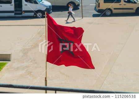 Recognizable national flag of Kingdom of Morocco sways in wind on ordinary day, when ordinary passers-by and cars move at a normal pace, confirming that despite dangers, life will go on Recognizable national flag of Kingdom of Morocco sways in wind on ordinary day, when ordinary passers-by and cars move at a normal pace, confirming that despite dangers, life will go on 115206538