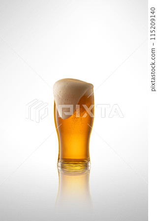 Tall glass filled with amber colored lager beer, topped with creamy white head against white studio background. 115209140