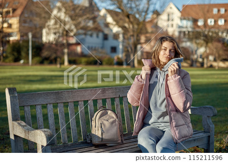 Winter Joy in Bitigheim-Bissingen: Beautiful Girl in Pink Jacket Sitting Amidst Half-Timbered Charm. beautiful girl in a pink winter jacket sitting on a bench in a park, set against the backdrop of 115211596