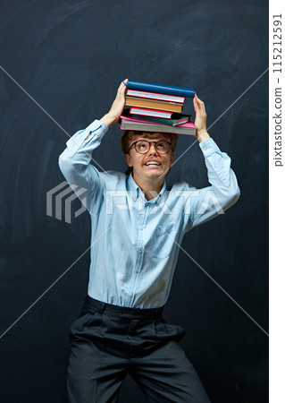 Emotional young redhaired man in formal attire trying holding stack of books above head against writing board background. 115212591