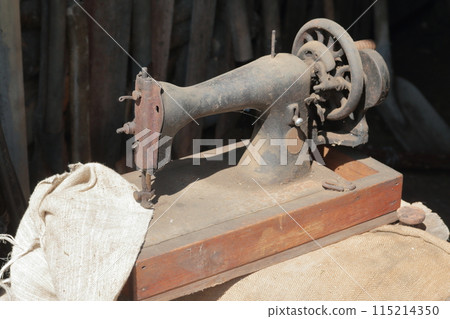 Old sewing machine, with manual drive. Covered with corrosion, dust and cobwebs. A piece of coarse fabric is sewn into it. Old sewing machine, with manual drive. Covered with corrosion, dust and cobwebs. A piece of coarse fabric is sewn into it. 115214350