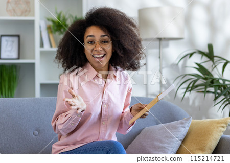 Happy young woman receiving and opening a mail envelope while sitting on a cozy sofa in a bright, modern living room. Happy young woman receiving and opening a mail envelope while sitting on a cozy sofa in a bright, modern living room. 115214721