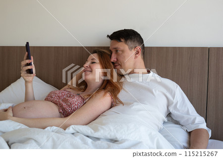 A happy couple relaxing in bed, sharing a joyful moment and looking at a smartphone together A happy couple relaxing in bed, sharing a joyful moment and looking at a smartphone together 115215267