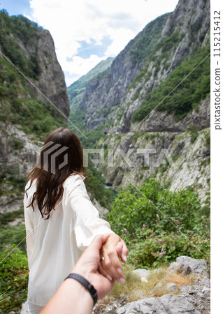 A girl stands with her back turned and a man holds her hand against the backdrop of a mountain view with lush green rocks under a cloudy sky. 115215412