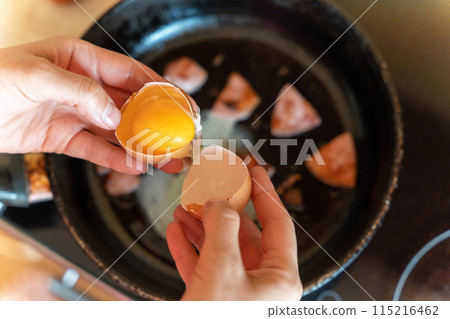 A hand cracks an egg over a frying pan of sizzling sausage slices 115216462