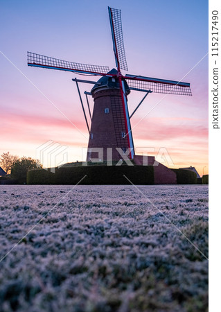 At sunset, a windmill stands in the middle of a field surrounded by nature At sunset, a windmill stands in the middle of a field surrounded by nature 115217490