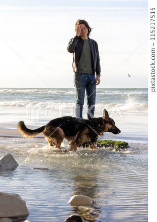 A man and a German Shepherd walk leisurely on the sunny beach, enjoying the serene atmosphere A man and a German Shepherd walk leisurely on the sunny beach, enjoying the serene atmosphere 115217515