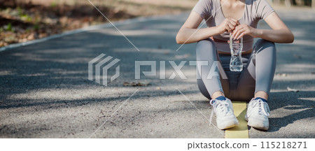 A woman sits on the ground with a bottle of water in her hand. She is wearing a grey shirt and grey pants A woman sits on the ground with a bottle of water in her hand. She is wearing a grey shirt and grey pants 115218271