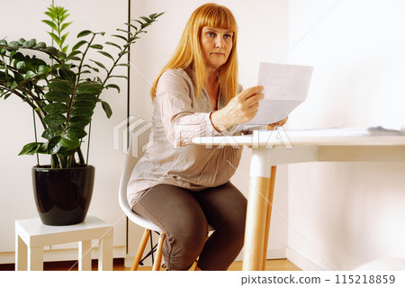 blonde woman reading letter notice while sitting at table in room 115218859