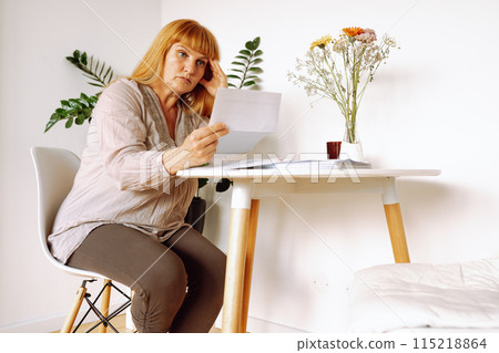blonde woman reading letter notice while sitting at table in room 115218864