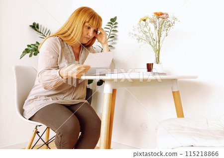 blonde woman reading letter notice while sitting at table in room blonde woman reading letter notice while sitting at table in room 115218866