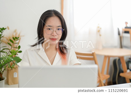 A young Asian woman wearing eyeglasses is working remotely from a cafe, working on her laptop. A young Asian woman wearing eyeglasses is working remotely from a cafe, working on her laptop. 115220691