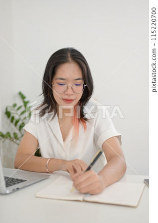 A focused Asian woman is working remotely from a cafe, taking notes or making list in her notebook A focused Asian woman is working remotely from a cafe, taking notes or making list in her notebook 115220700