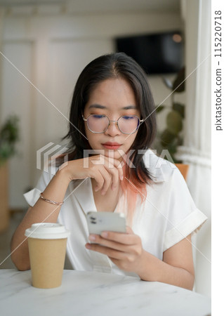 A woman is reading messages on her smartphone while sitting at a table by the window in a cafe. 115220718