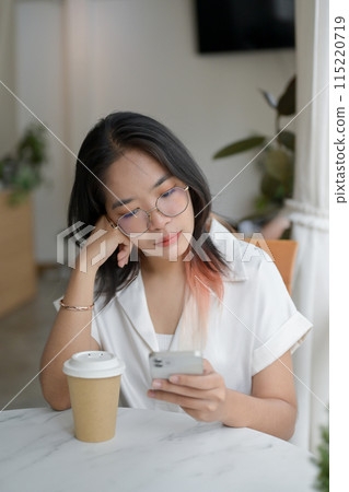 A woman is waiting for someone in the coffee shop, looking at her smartphone with boredom. 115220719
