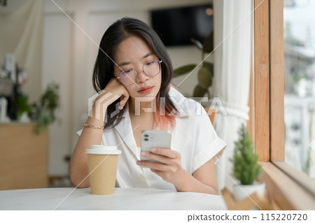 A woman is waiting for someone in the coffee shop, looking at her smartphone with boredom. 115220720