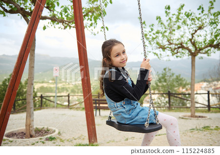 Little child girl smiling and dreamily looking away while sitting on a wooden swing in the playground outdoors. People. Happy carefree childhood. Recreation. Kids development and entertainment 115224885