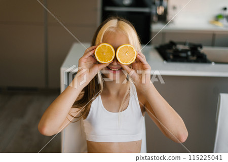 Smiling child girl covered his eyes with orange halves sitting at the table in the kitchen. Citrus fruits for child food Smiling child girl covered his eyes with orange halves sitting at the table in the kitchen. Citrus fruits for child food 115225041