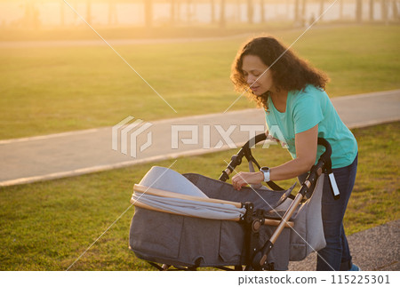 Smiling happy young woman, loving caring mother walking with her newborn baby in stroller, spending time funny on the city park t sunset Smiling happy young woman, loving caring mother walking with her newborn baby in stroller, spending time funny on the city park t sunset 115225301
