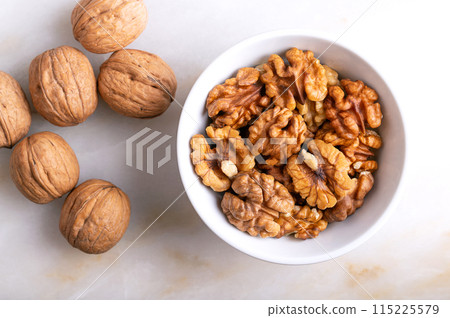 Walnuts in a white bowl on a marble plate. Shelled and dried halves of walnut kernels, ripe seeds of the common walnut tree Juglans regia, used as snack or for baking. Close-up from above, food photo. 115225579