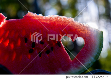 Man holding red watermelon slice cut into heart shape in green garden background. Valentine's Day love concept. Sunset outdoors summer day. Healthy seasonal local food 115225596