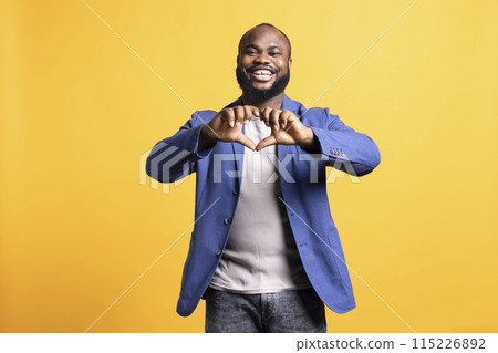 Portrait of loving joyous african american man doing heart symbol shape with hands. Happy caring BIPOC person showing love gesturing, isolated over yellow studio background Portrait of loving joyous african american man doing heart symbol shape with hands. Happy caring BIPOC person showing love gesturing, isolated over yellow studio background 115226892