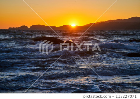 Choppy waves with boulders in water and golden sky with sun poking above mountains at sunset 115227071