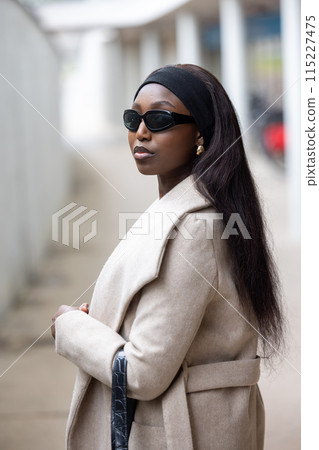 A stylish woman in a beige coat and sunglasses exudes confidence while being outdoors 115227475