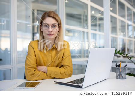 Confident businesswoman sitting at a desk in a modern office equipped with a laptop, tablet, and pens. Professional and focused atmosphere. 115229117
