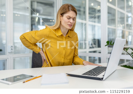 Woman holding her lower back in pain while working on a laptop at her desk in a modern office environment. 115229159