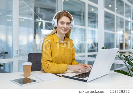 Smiling businesswoman wearing headphones working on a laptop in a modern office setting. Coffee cup and notebook are on the desk. Smiling businesswoman wearing headphones working on a laptop in a modern office setting. Coffee cup and notebook are on the desk. 115229232