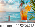 cocktail in glass with straw and ice on the table on the beach, ocean and palm leaf as background 115230818