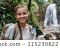 smiling young woman tourist with backpack holding bottle of water against the backdrop of mountain waterfall 115230822