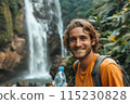 happy young man tourist with backpack holding bottle of water against the backdrop of mountain waterfall 115230828