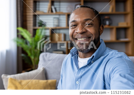 A cheerful man with a beard and blue shirt smiling warmly while sitting on a cozy couch in a modern living room 115231082