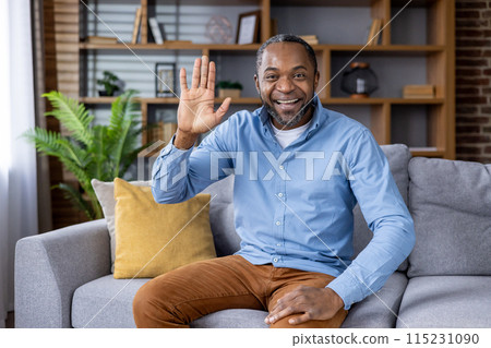 Smiling man waving while sitting on a comfortable sofa in a cozy, stylish living room with modern decor. Smiling man waving while sitting on a comfortable sofa in a cozy, stylish living room with modern decor. 115231090