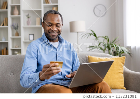 Smiling man holding a credit card and using a laptop while shopping online at home. Comfortable and modern living environment Smiling man holding a credit card and using a laptop while shopping online at home. Comfortable and modern living environment 115231091