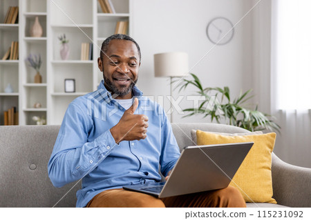 Smiling man sitting on the couch at home, giving a thumbs up gesture while using a laptop for work or communication. 115231092
