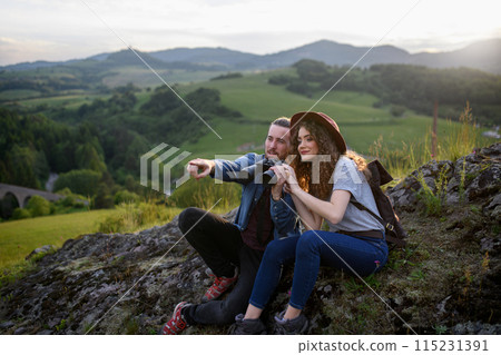Travellers couple hiking on easy trail in nature with backpacks. Young tourist spending summer vacation oudoors, sitting on rocks and enjoying breathtaking view. Travellers couple hiking on easy trail in nature with backpacks. Young tourist spending summer vacation oudoors, sitting on rocks and enjoying breathtaking view. 115231391