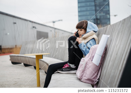 Sad girl phone calling, sitting in front of school building, waiting for parents. Young schoolgirl with no friends, feeling lonely. 115231572