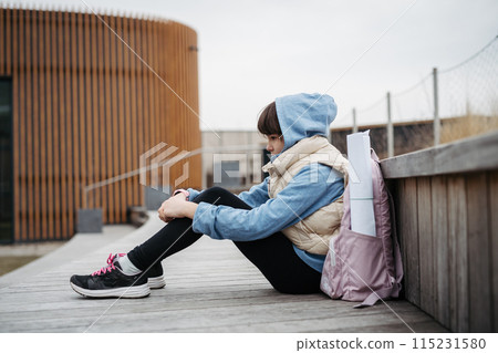 Sad girl alone, sitting in front of school building, waiting for parents. Young schoolgirl with no friends, feeling lonely. 115231580