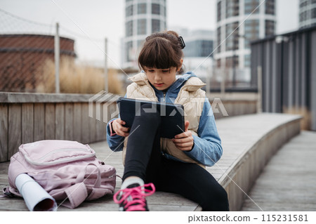 Girl sitting in front of school building, scrolling on tablet. Using social media. Online bullying or cyberbullying for young schoolgirl. Girl sitting in front of school building, scrolling on tablet. Using social media. Online bullying or cyberbullying for young schoolgirl. 115231581