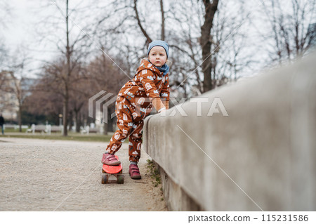 Cute toddler girl playing outdoor in playground with skateboard. Girl in softshell bodysuit spending time in park. 115231586