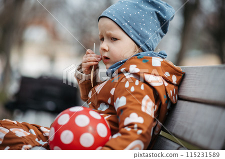 Cute toddler girl eating lunch in park, sitting on bench. Picnic in park during cold spring day. 115231589